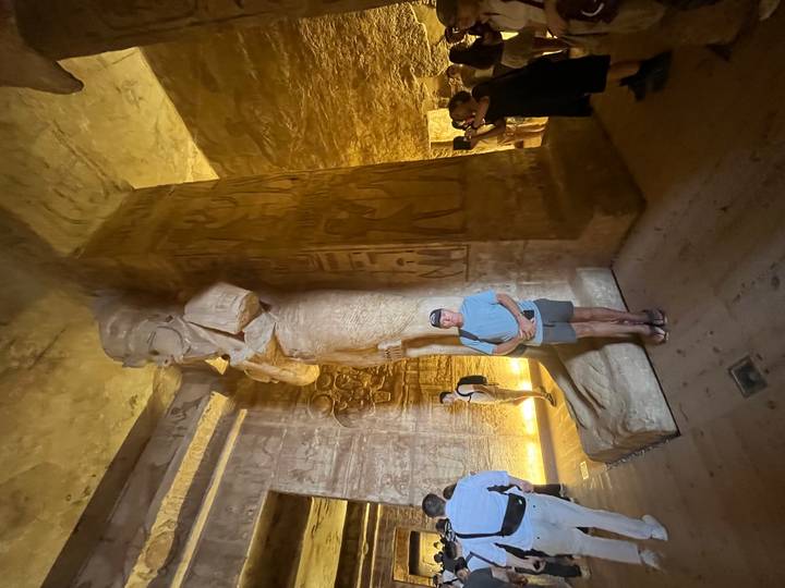 Traveller poses beside a colossal pharaonic statue inside a dimly lit rock-cut chamber with carved walls.