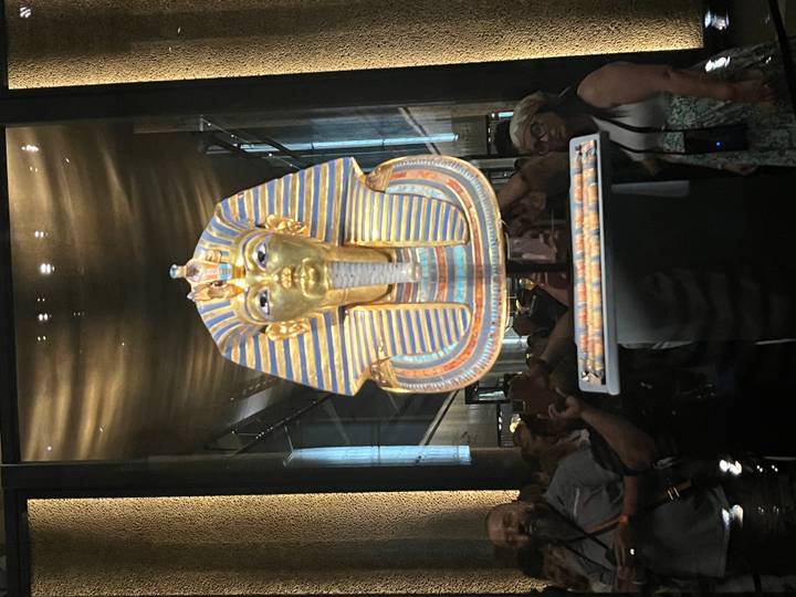 The gold funerary mask of Tutankhamun displayed behind glass, surrounded by a crowd taking photos.