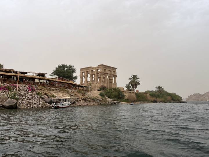 Ancient stone temple of Philae rises beside the Nile, viewed from a boat on choppy water under hazy skies.