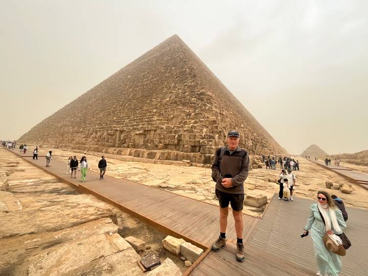 Traveller poses before the Great Pyramid of Giza on a hazy day while visitors walk the surrounding boardwalk.