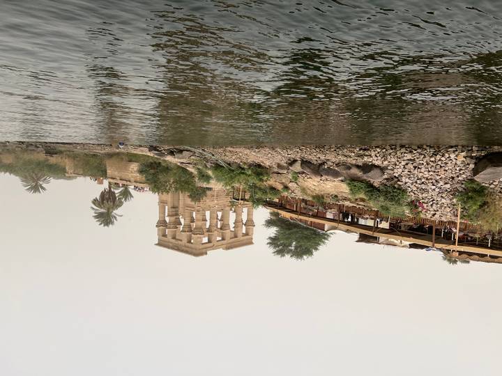 View from the water of the ancient Kiosk of Trajan on a rocky riverbank with palms.
