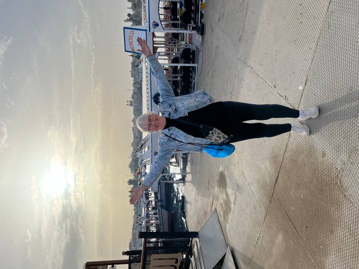 A smiling woman raises her arms on a pier with boats and sunrise light in the background.
