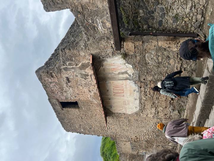 Tourists walking past a weathered stone wall with faded Roman frescoes in Pompeii on an overcast day.