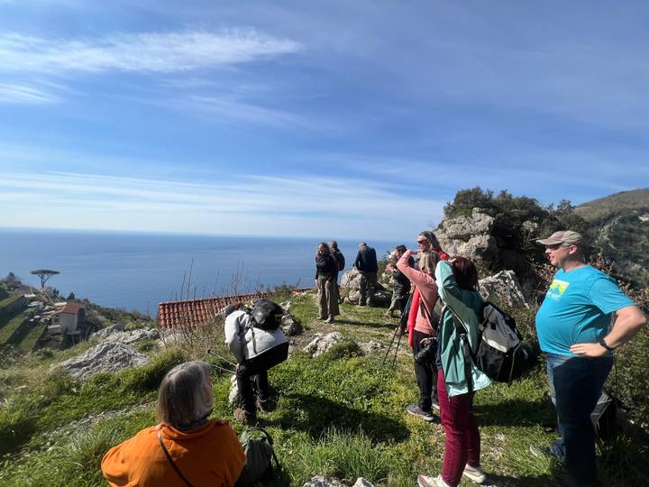 Hikers pausing at a cliff-top viewpoint overlooking the Mediterranean Sea and blue sky.