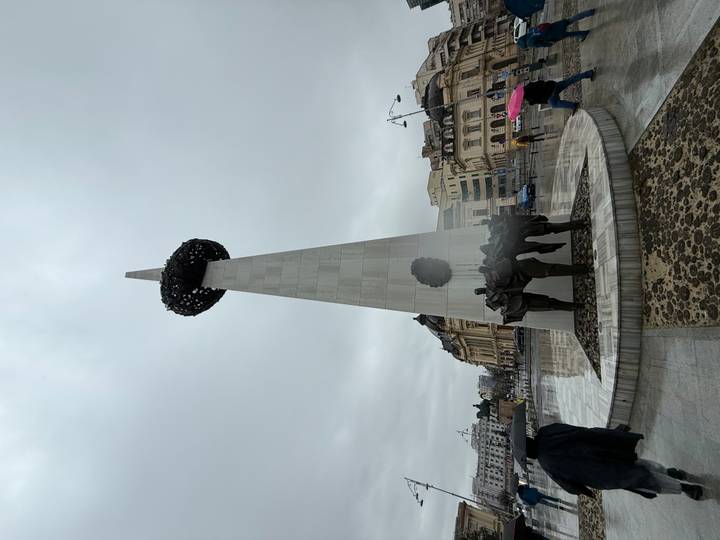 Tall modern marble obelisk with a wreath and bronze statues at its base in a city square on a gray overcast day.