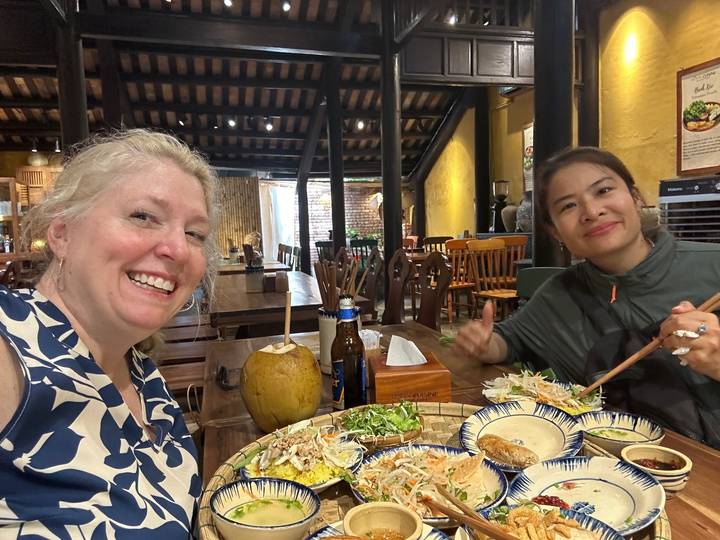 Two women enjoying a Vietnamese feast at a wooden restaurant table, smiling at the camera.