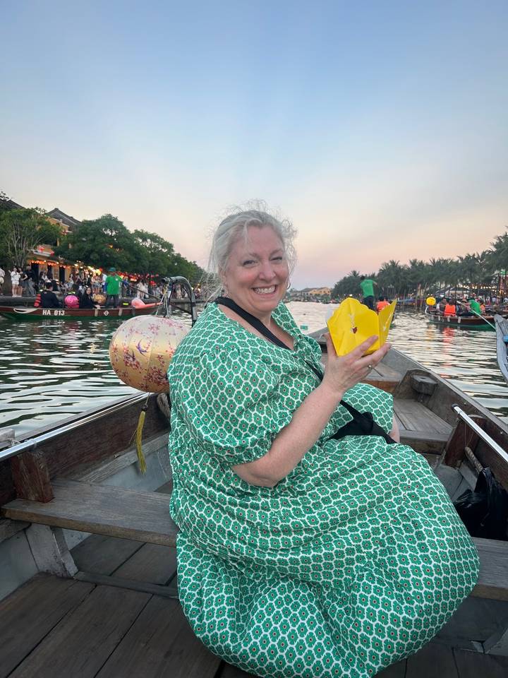 Smiling woman in green dress holding a yellow lantern flower while sitting in a small boat on a lantern-lit river.