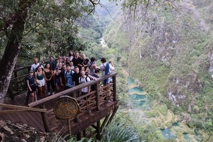 Large tour group posing on a wooden lookout deck above the turquoise pools of Semuc Champey surrounded by jungle cliffs.