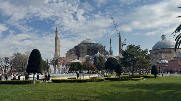 Wide garden view of Hagia Sophia with fountains, domes and cranes under patchy clouds.