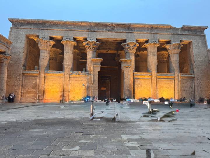 Facade of an ancient Egyptian temple lit warmly at dusk with blurred visitors walking across the courtyard