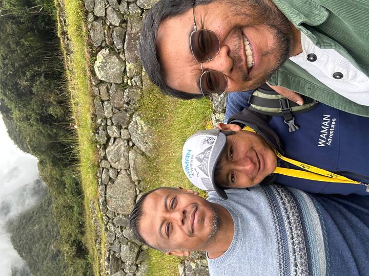 Three friends take a close selfie with stone terraces and misty mountains in the background.