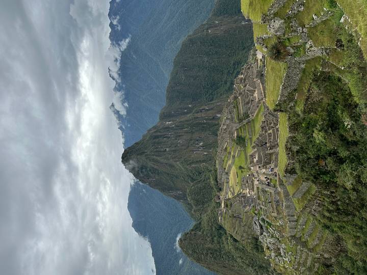 Wide view of Machu Picchu citadel nestled below a steep green peak under overcast skies.