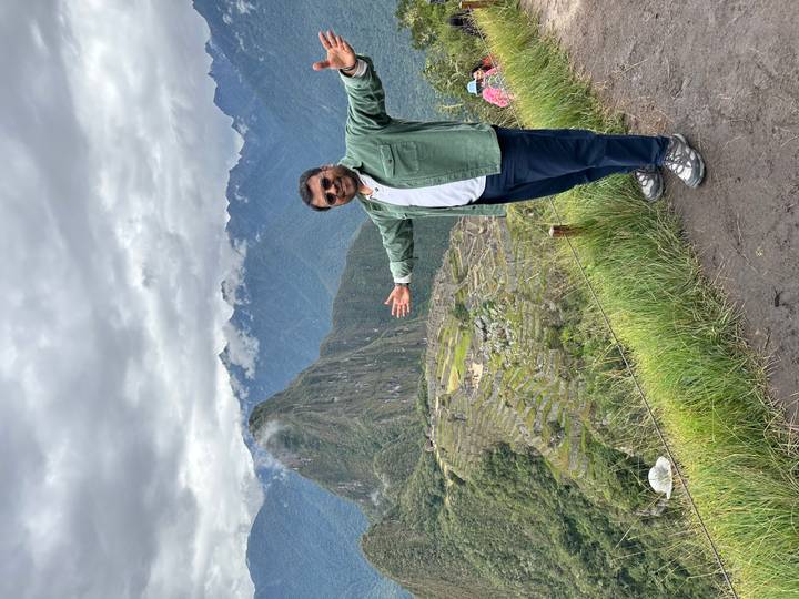 A traveler poses joyfully on a ledge overlooking Machu Picchu and cloud-capped Andes peaks.