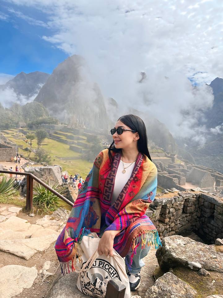 A woman wrapped in a colorful shawl looks over misty Machu Picchu ruins partially veiled by clouds.