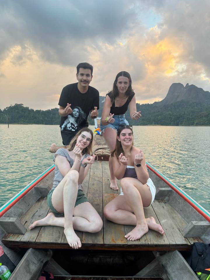 Friends make heart gestures while sitting on the prow of a long-tail boat on a lake at sunset.