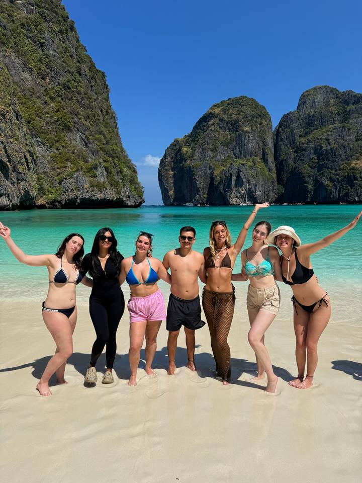 Group of travelers in swimwear celebrate on a pristine tropical beach with turquoise water and limestone cliffs.