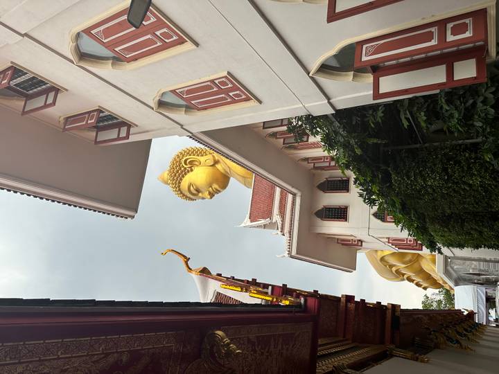 Giant golden Buddha head rises above temple rooftops framed by white walls and red shutters.