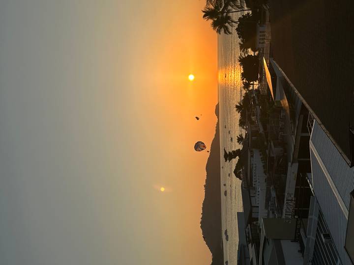 Orange sun sets over the sea with parasailers gliding against the glowing sky.