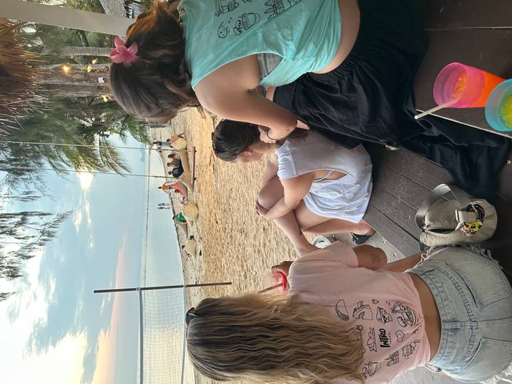 Young travelers relax on a wooden deck by a sandy beach, sipping colorful cocktails as the sun lowers over calm sea waters with a volleyball net nearby.