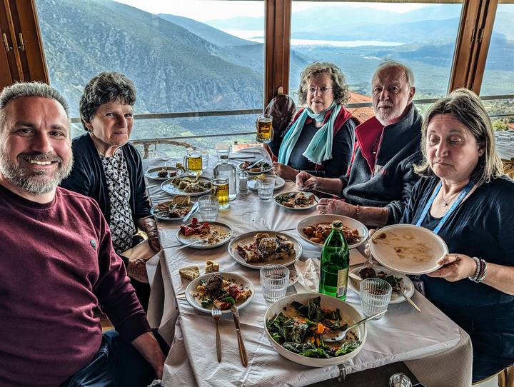 Smiling travellers enjoy a hearty Greek meal around a table with panoramic hillside views beyond the window.