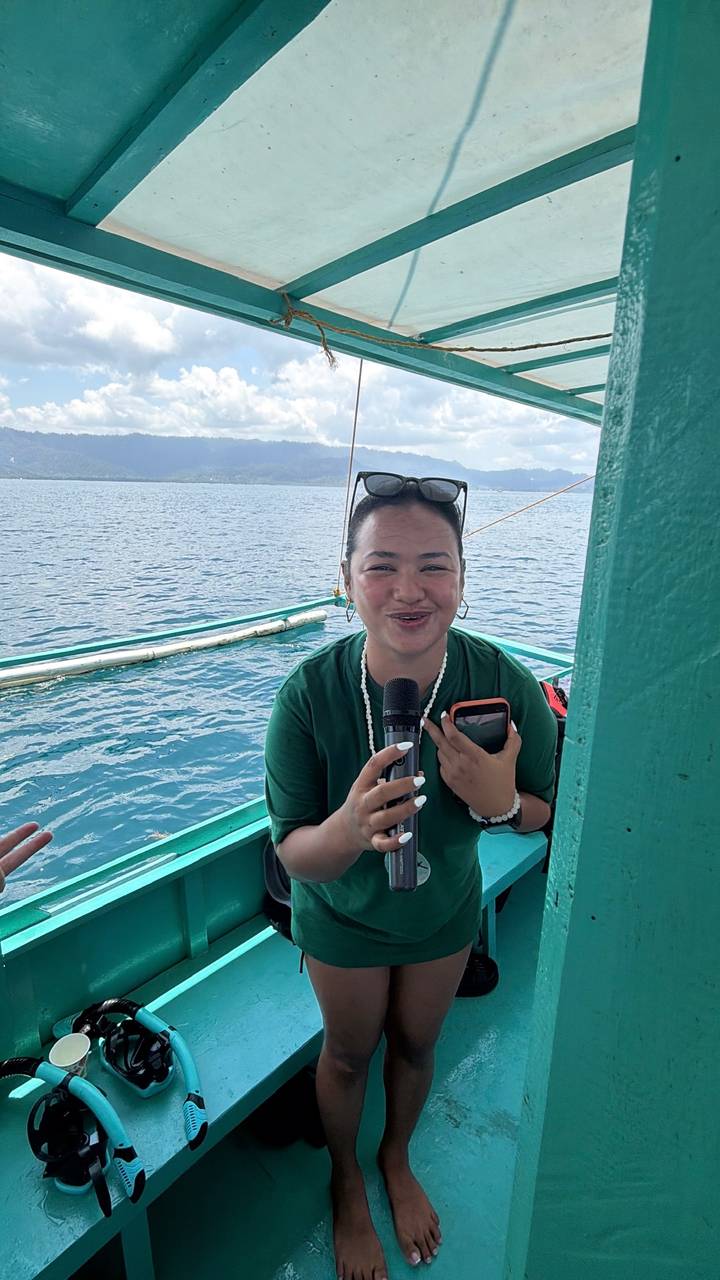 Smiling woman on a boat holding a microphone and phone with turquoise sea and islands in the background