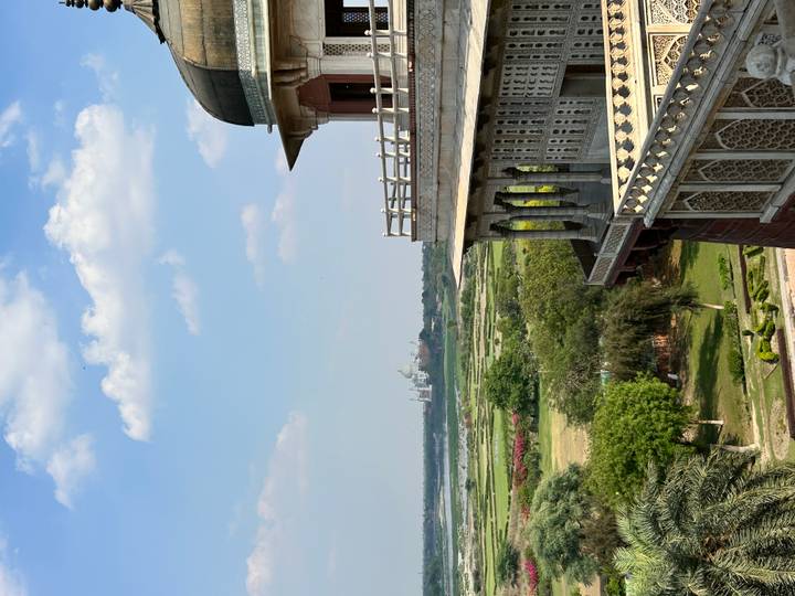 Scenic view from a historic fort balcony looking across green plains and the distant Taj Mahal under a blue sky with scattered clouds