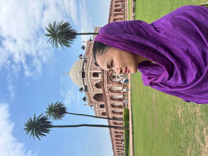 Close-up of a woman in a purple scarf with Humayun's Tomb and palm trees in the background against a bright blue sky
