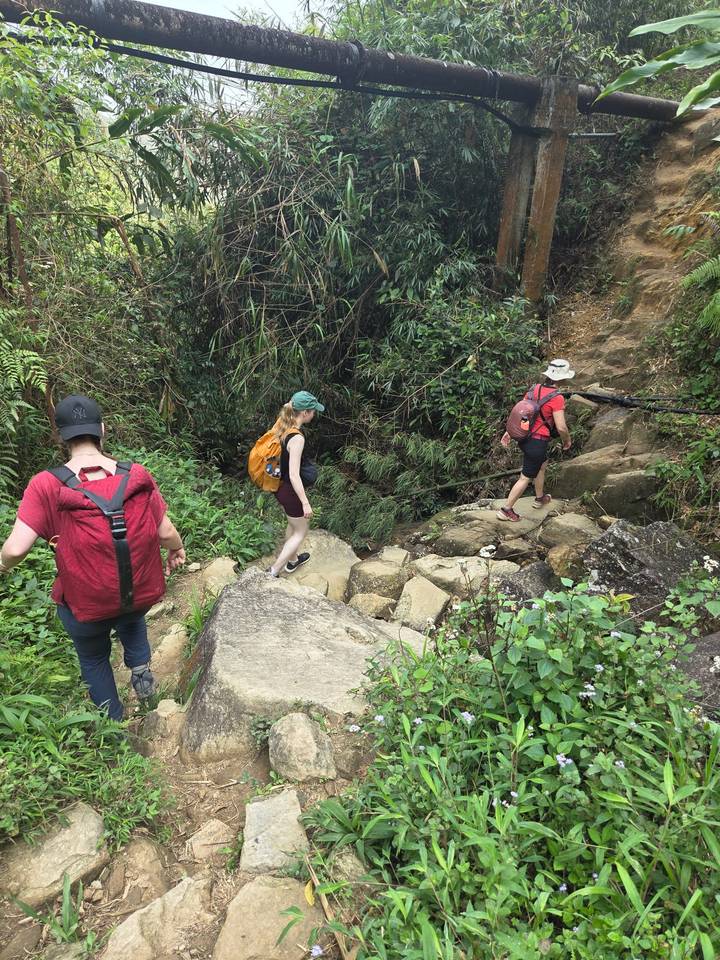 Three hikers with backpacks carefully crossing rocky terrain surrounded by dense jungle vegetation