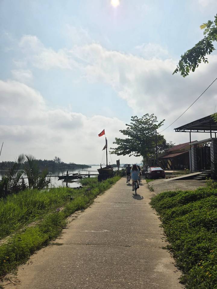 Cyclists riding along a riverside path beneath a Vietnamese flag with boats and lush greenery nearby