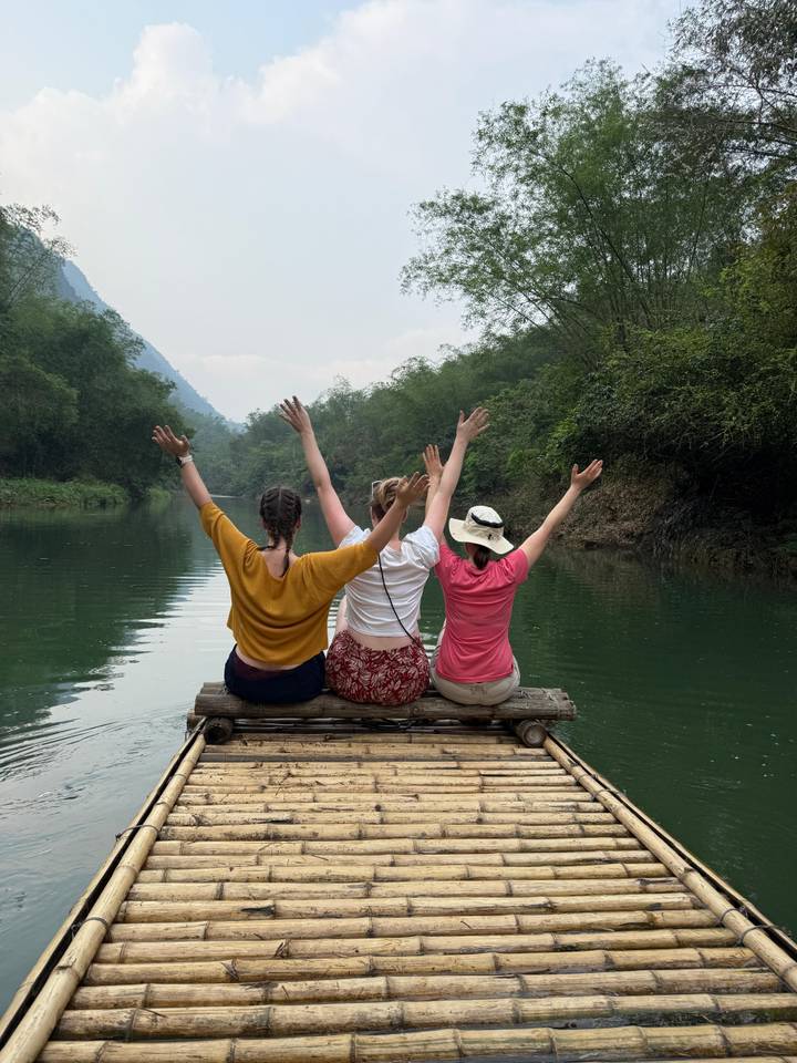 Three women seated on a bamboo raft raising their arms joyfully while floating through a forested river canyon