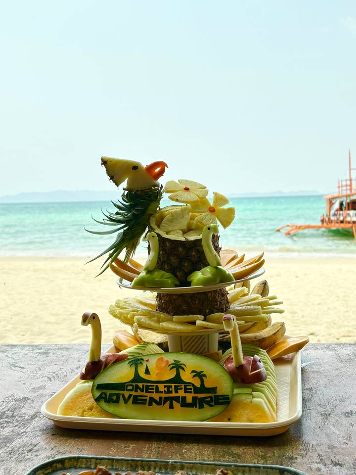 Elaborate tropical fruit carving platter displayed on sandy beach with turquoise sea behind