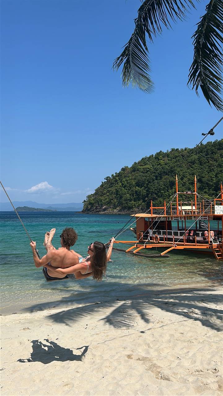 Couple on over-water swing enjoying clear blue lagoon with tour boat nearby