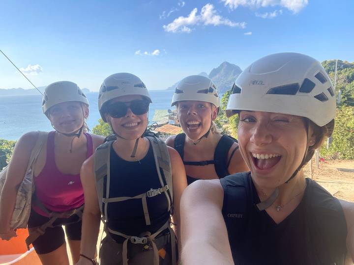 Smiling group selfie of four women wearing climbing helmets with coastal mountains behind