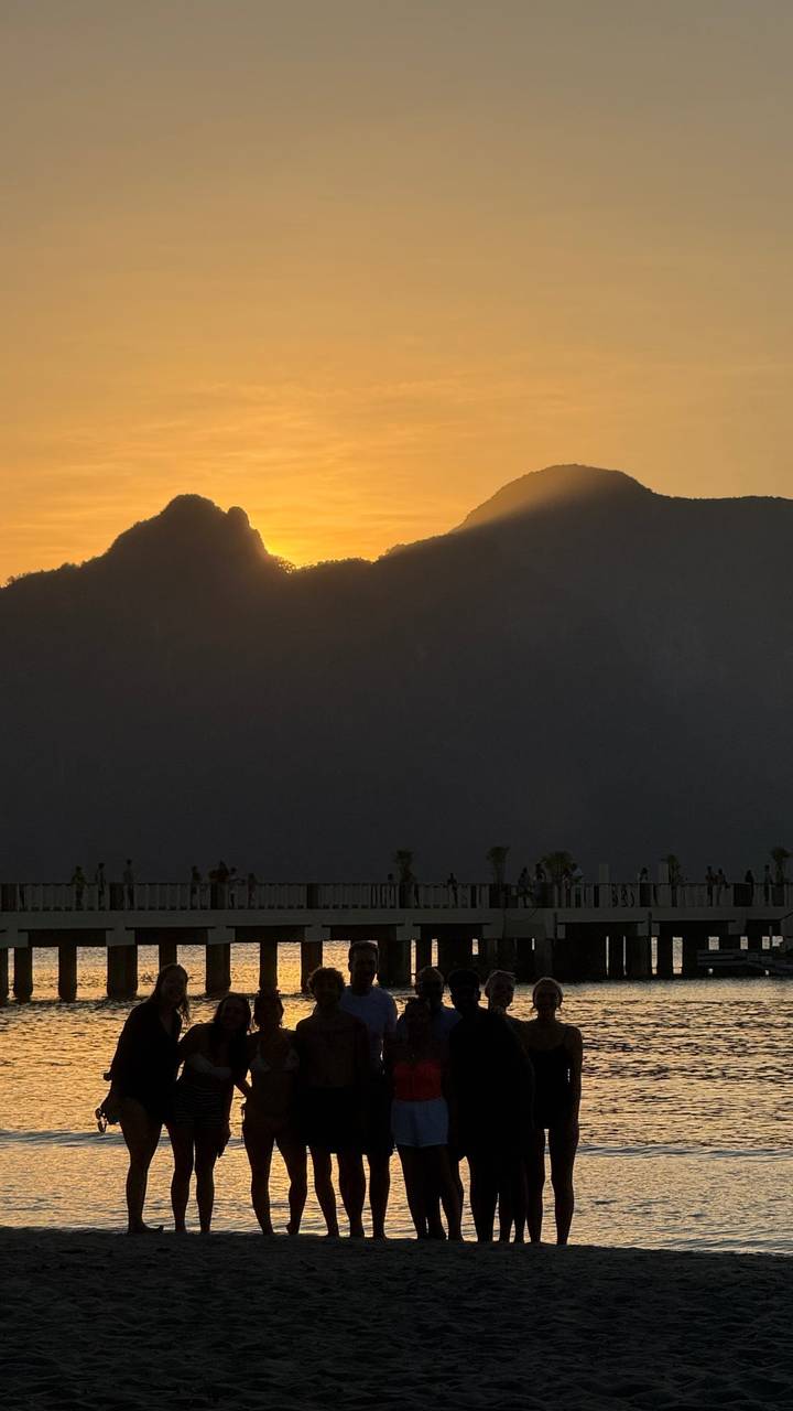 Silhouetted visitors on coastal boardwalk watching dramatic orange sunset behind mountains