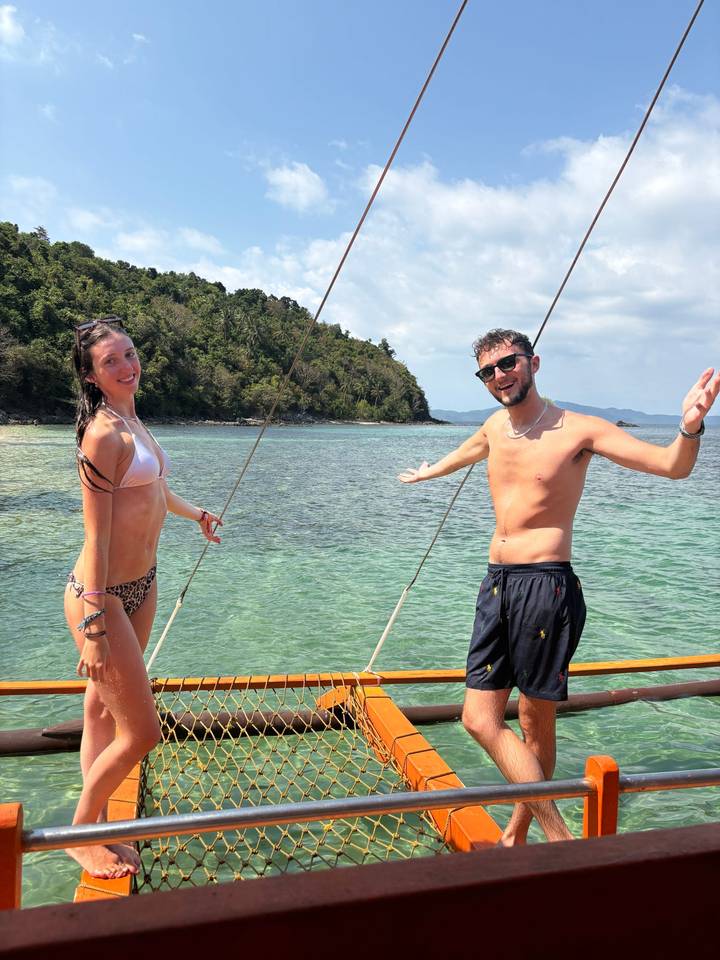 Two smiling travellers in swimwear pose on a sailboat with turquoise tropical water and forested islands behind them.