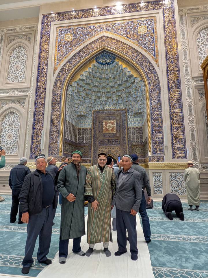 Four men wearing traditional caps stand before an intricately tiled mosque interior.