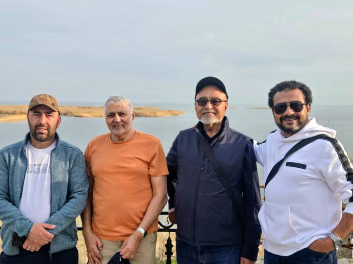 Four men pose on a balcony overlooking the calm waters and barren shoreline of a large lake.