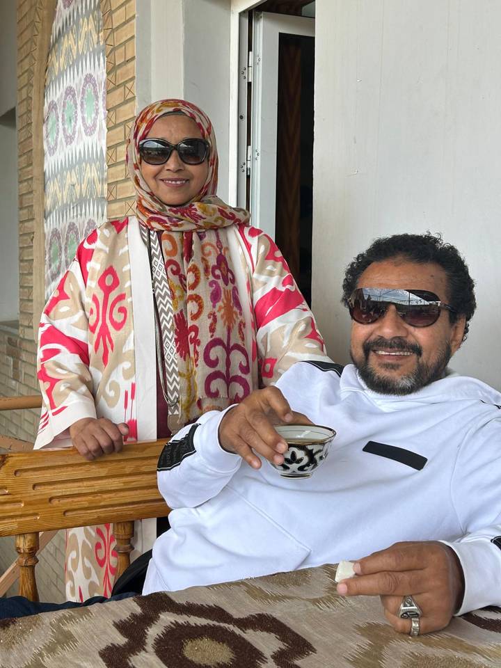 A man wearing sunglasses enjoys a cup of tea while a woman stands beside him on a balcony.