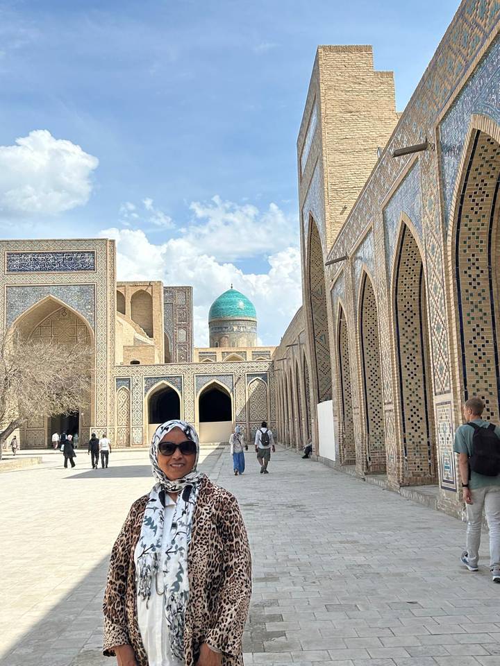 A woman smiles in a courtyard framed by tiled arches and a turquoise dome in the distance.