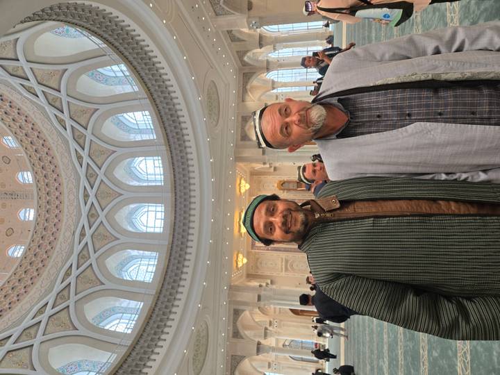 Two men in traditional attire stand under a grand white marble dome with patterned ceiling.