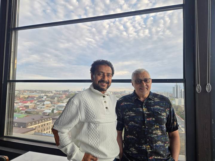 Two men pose by a large window overlooking a sprawling cityscape at dusk.