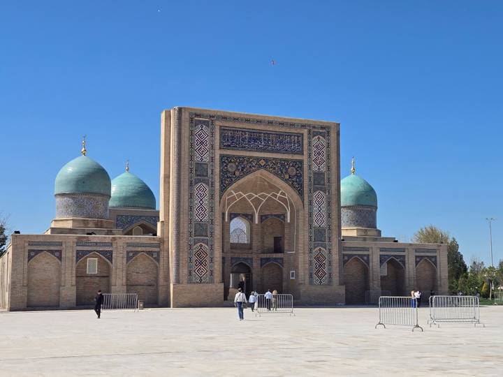 The Hazrati Imam complex with three turquoise domes and intricate portal stands under a clear blue sky.