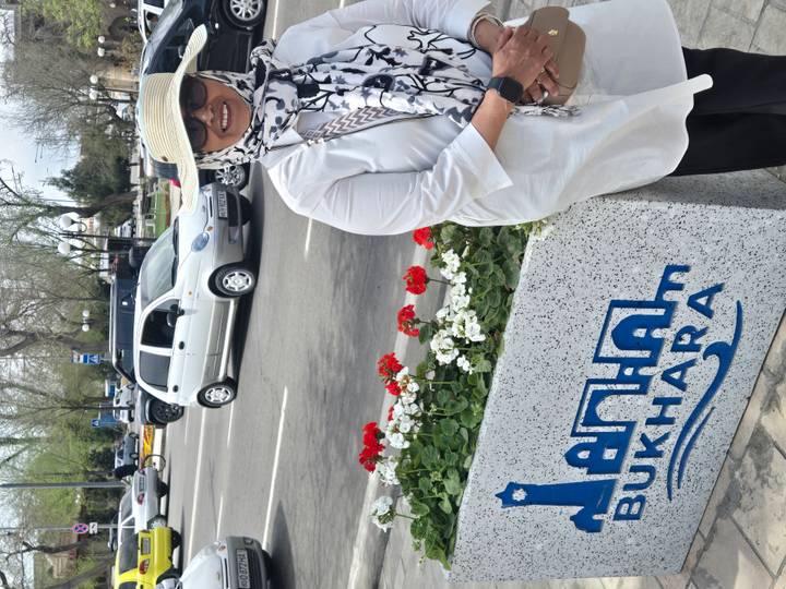 A woman in sunglasses smiles beside a flower bed and a decorative Bukhara city sign.