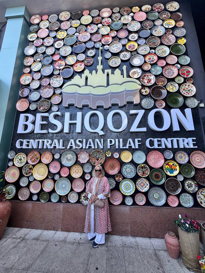 A woman stands beneath a large sign for the Central Asian Pilaf Centre decorated with colorful plates.