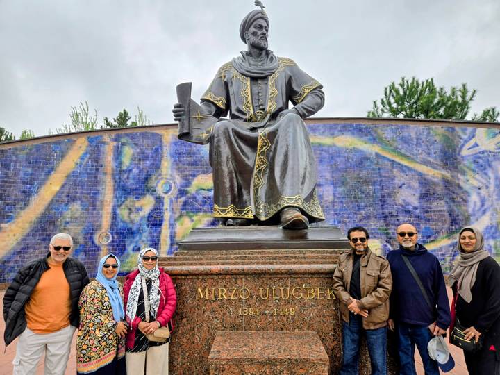 Travellers pose in front of the seated bronze statue of Ulugbek with a colorful mural backdrop.