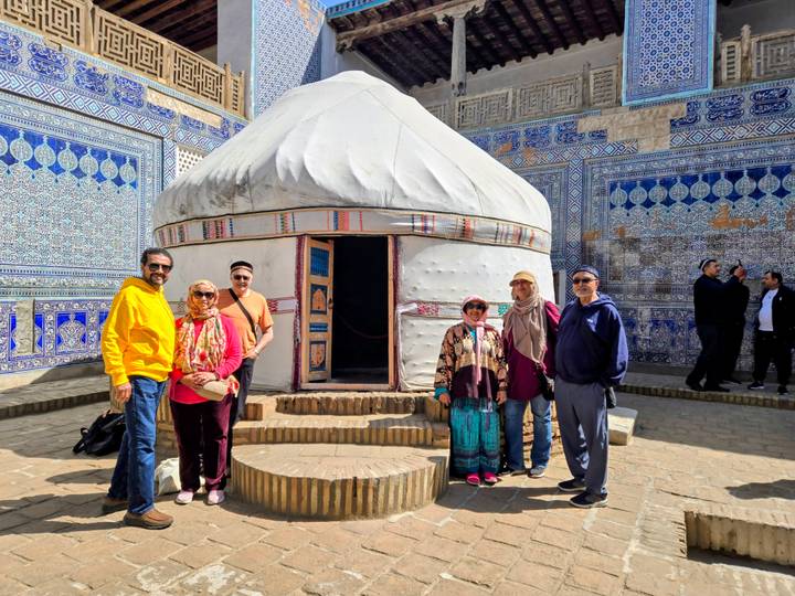 Travellers stand beside a traditional yurt set among ornate blue tile walls.