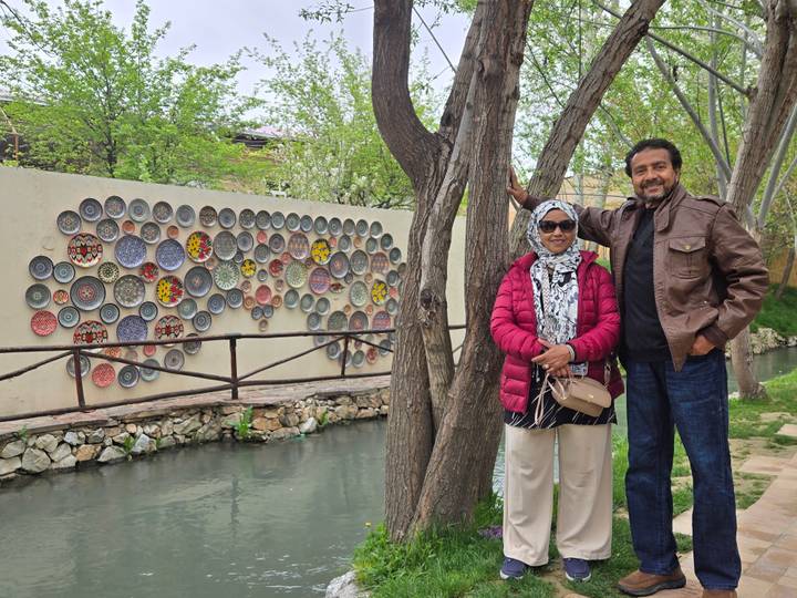 A couple poses beside a decorative wall of colorful ceramic plates next to a narrow canal and trees.