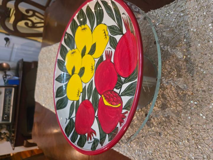 A brightly painted ceramic plate featuring red pomegranates and yellow flowers on a glass stand indoors.