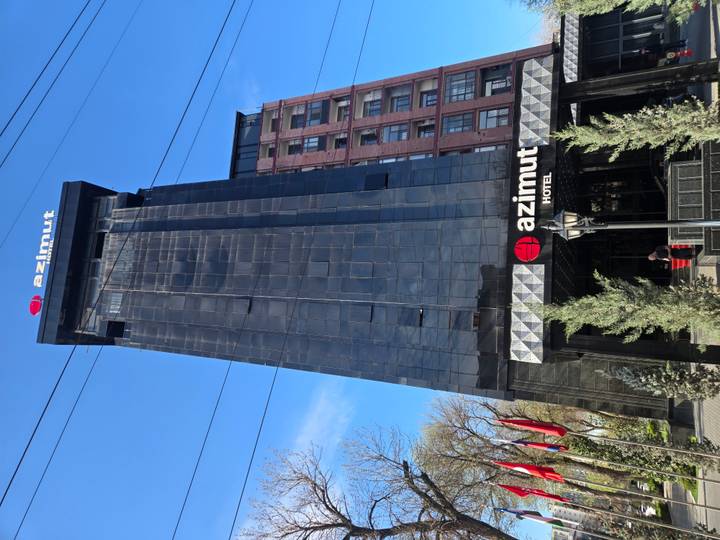 Tall black glass façade of the Azimut Hotel rises against a clear sky with flags at its entrance.