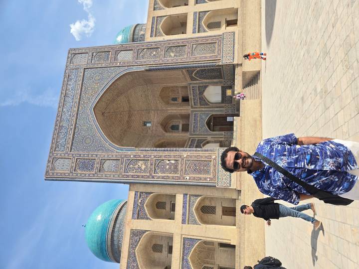 A smiling man stands before a grand tiled portal with turquoise domes under a bright sky.
